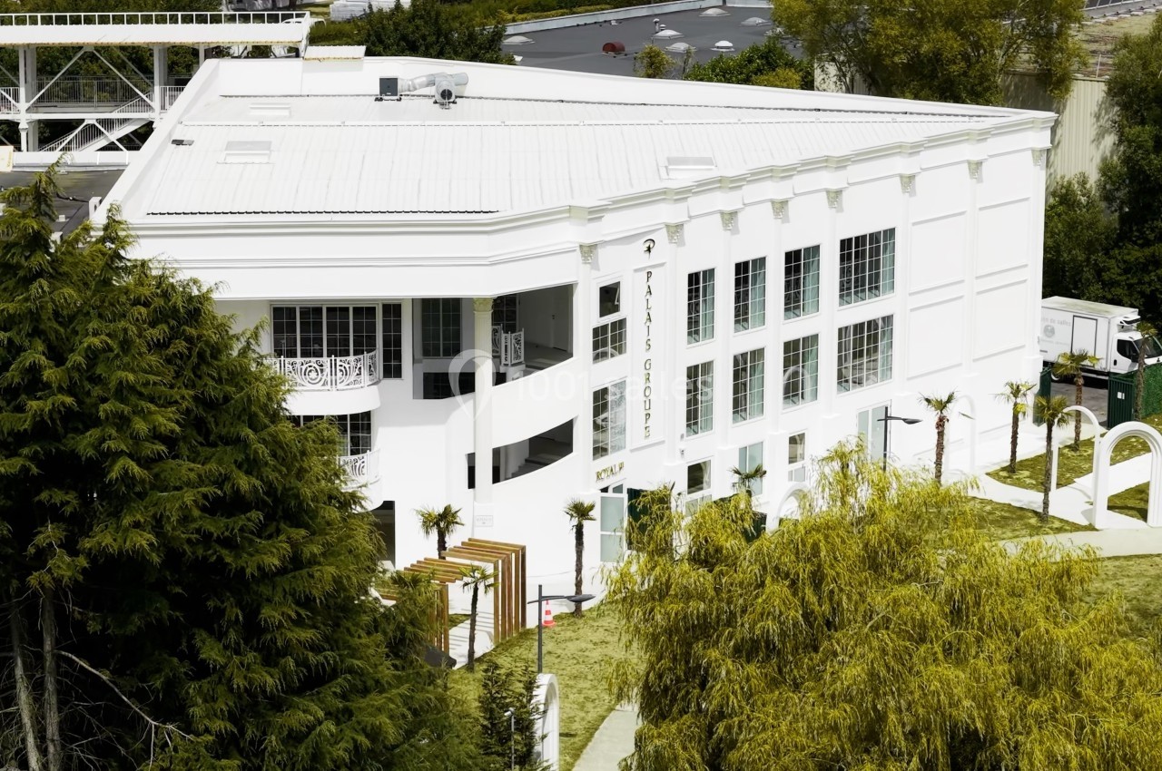 Bâtiment blanc moderne entouré d'arbres et de verdure, avec un parking visible en arrière-plan.