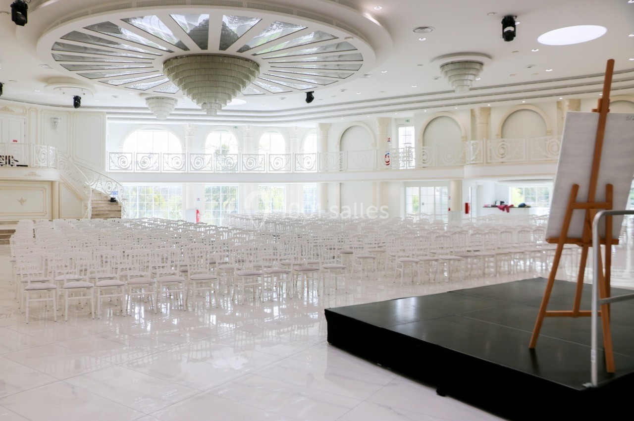 Salle de réception lumineuse avec des chaises blanches alignées, un podium noir et un chevalet au premier plan.