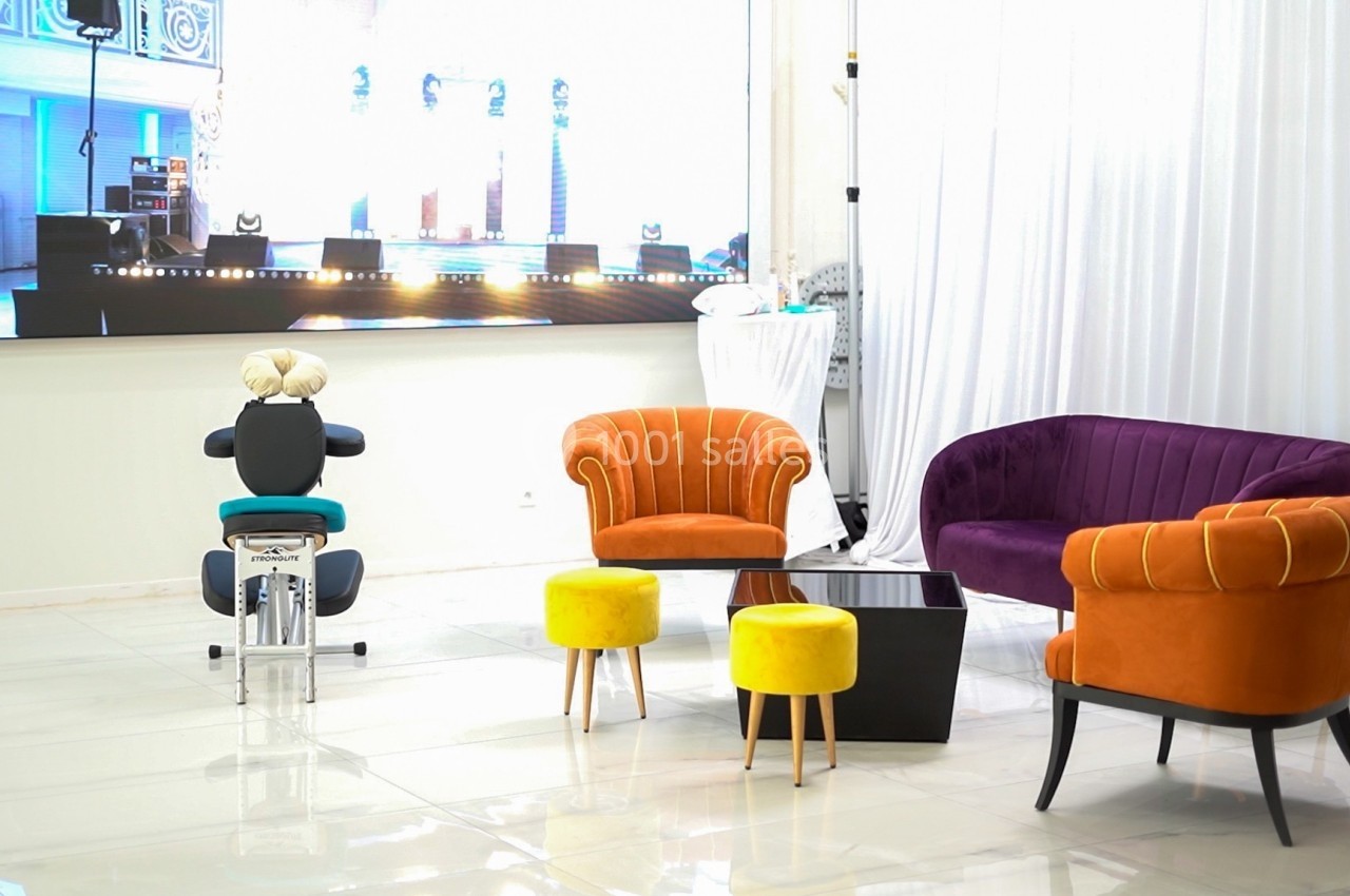 Salon moderne avec fauteuils colorés, tabourets jaunes, table basse noire et écran lumineux en arrière-plan.