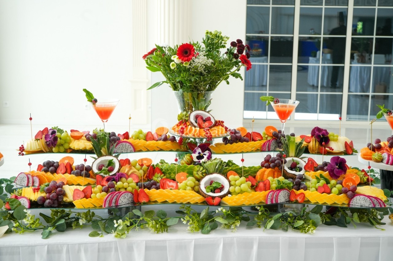 Buffet décoratif avec des fruits variés, disposés en étages, entouré de fleurs et de cocktails colorés.