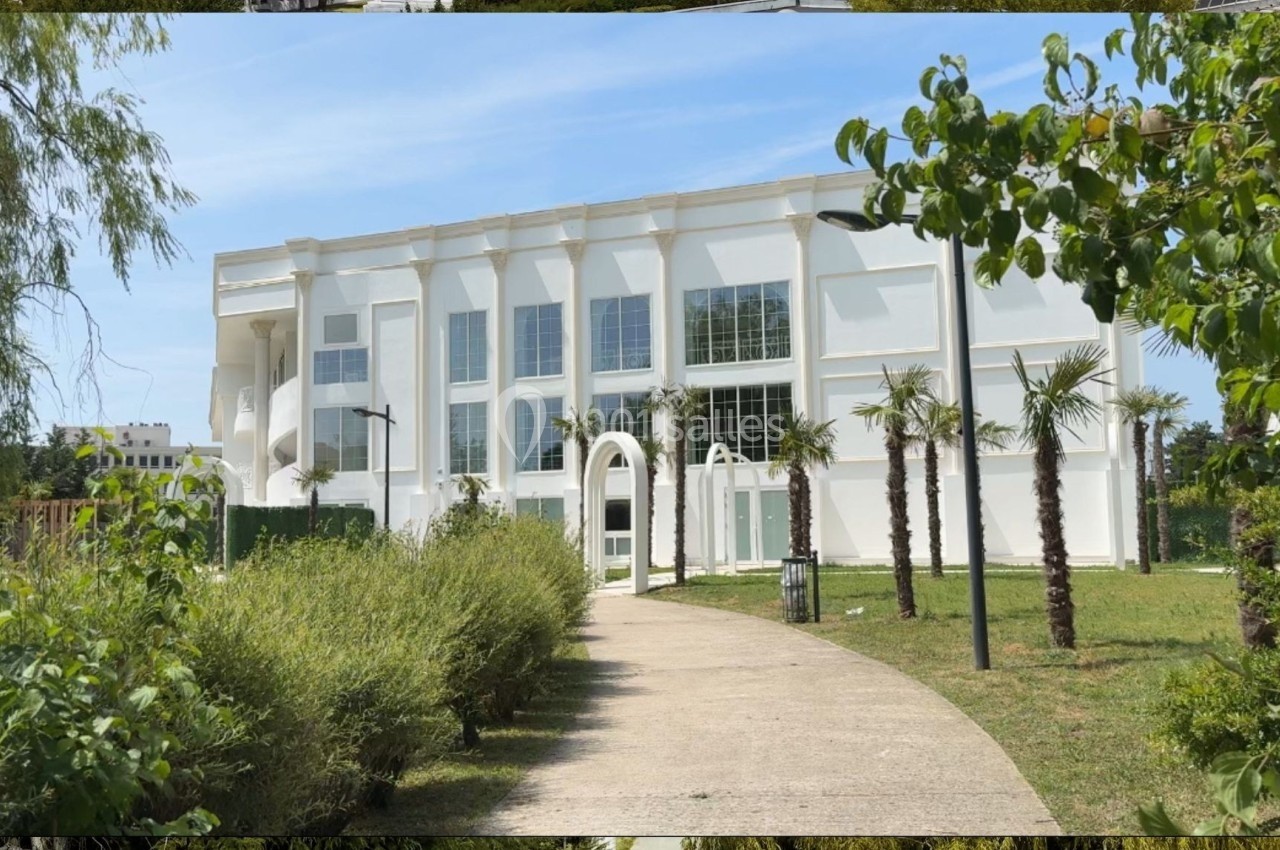 Bâtiment blanc moderne entouré de végétation, avec un chemin pavé menant à une entrée voûtée.