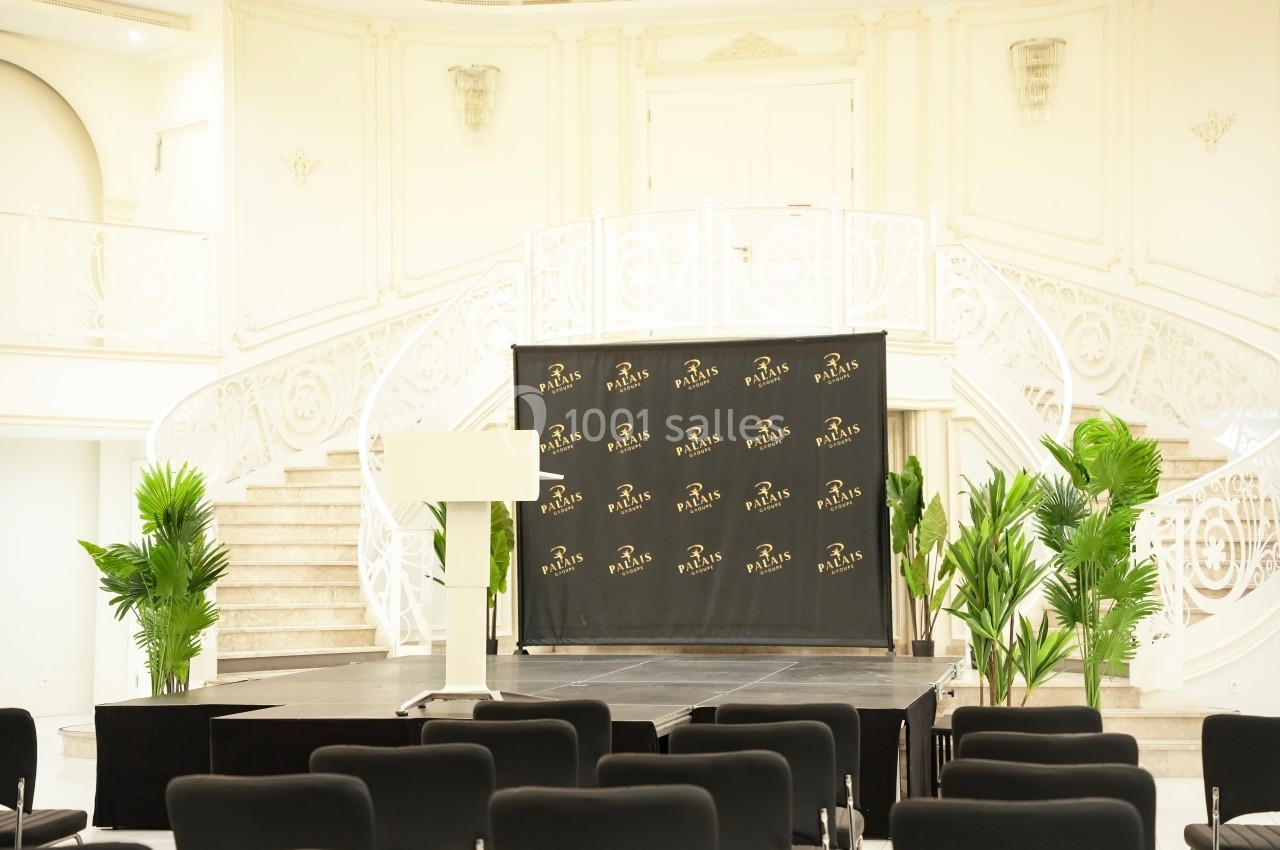 Salle de conférence lumineuse avec estrade, pupitre, chaises noires et escalier orné de balustrades blanches.