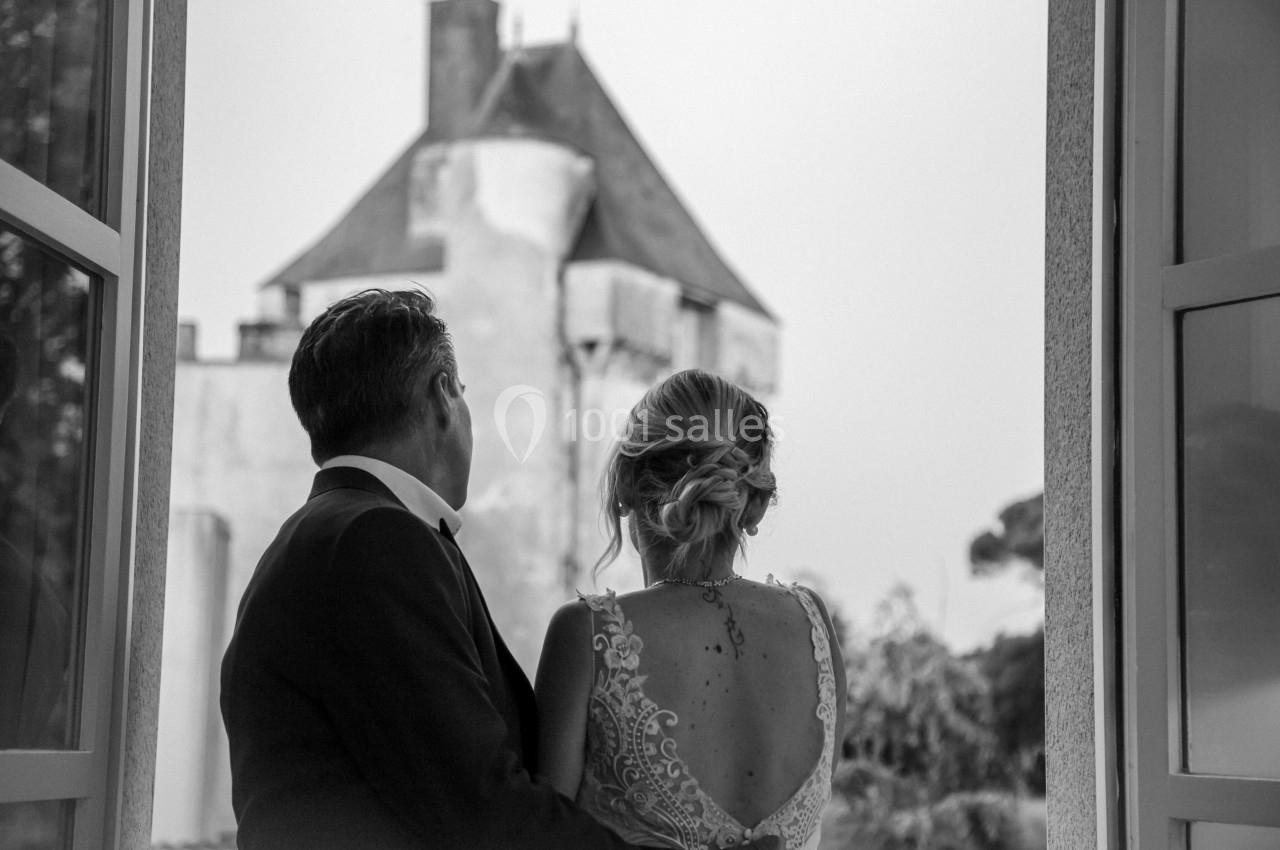 Un couple de dos regarde par une fenêtre ouverte un bâtiment ancien avec une tour, en noir et blanc.