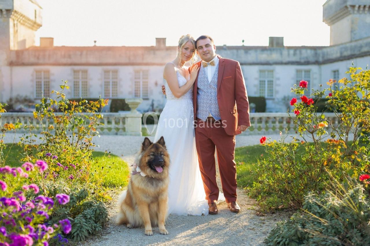Un couple en tenue de mariage pose avec un chien dans un jardin fleuri devant un bâtiment ancien.