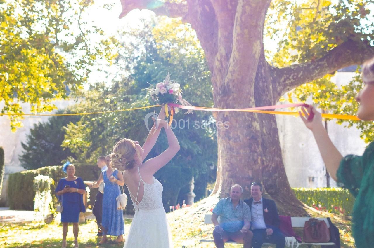 Une mariée lance son bouquet dans un jardin ensoleillé, entourée d'invités sous un grand arbre.