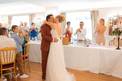 Un couple de mariés s'embrasse devant des invités applaudissant dans une salle de réception lumineuse.
