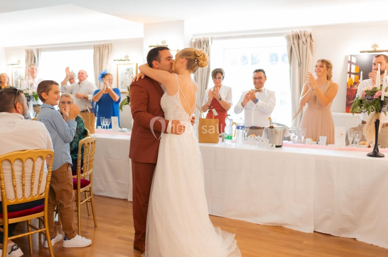 Un couple de mariés s'embrasse devant des invités applaudissant dans une salle de réception lumineuse.