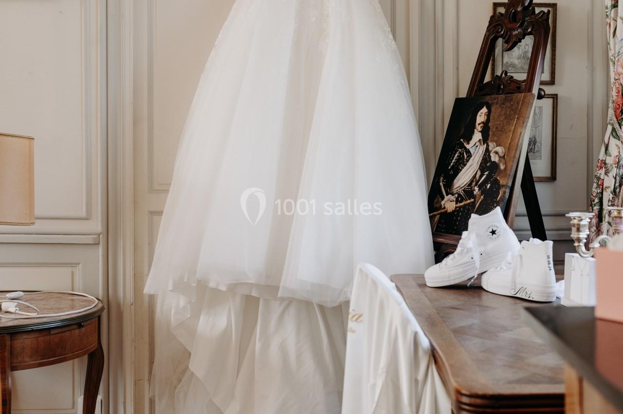 Robe de mariée blanche suspendue à une porte dans une pièce élégante avec parquet et meubles anciens.