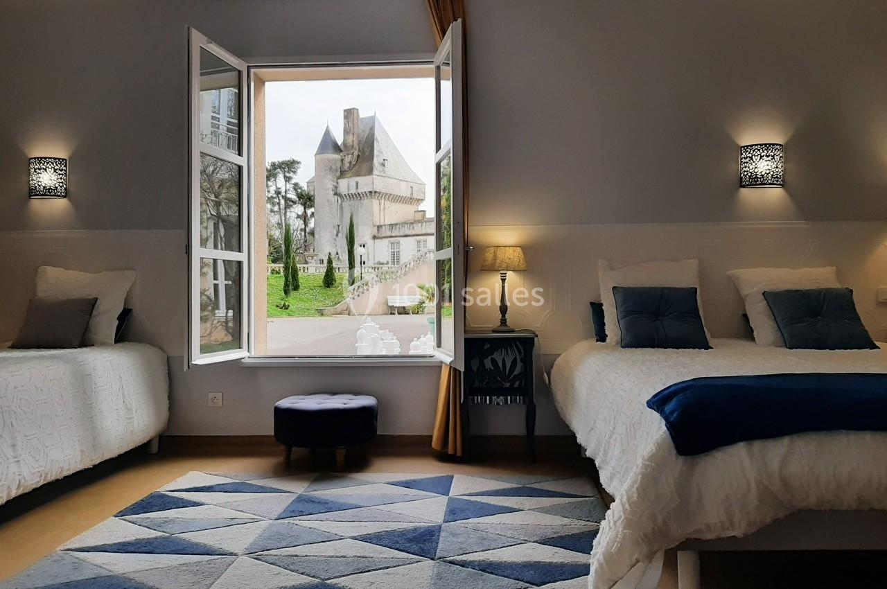 Chambre lumineuse avec deux lits, fenêtre ouverte offrant une vue sur un château entouré de verdure.