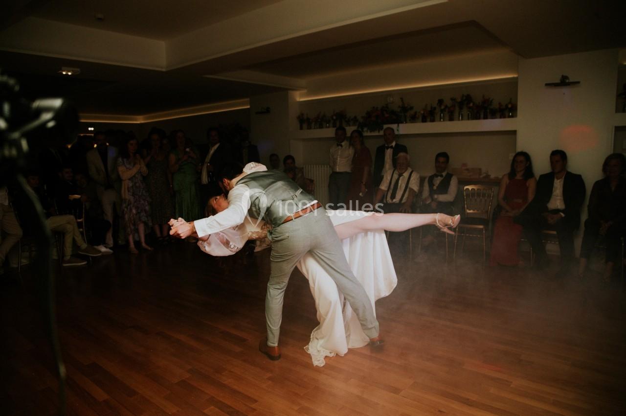 Un couple effectue une danse de mariage avec une figure élégante sur une piste en bois, entouré d'invités attentifs.