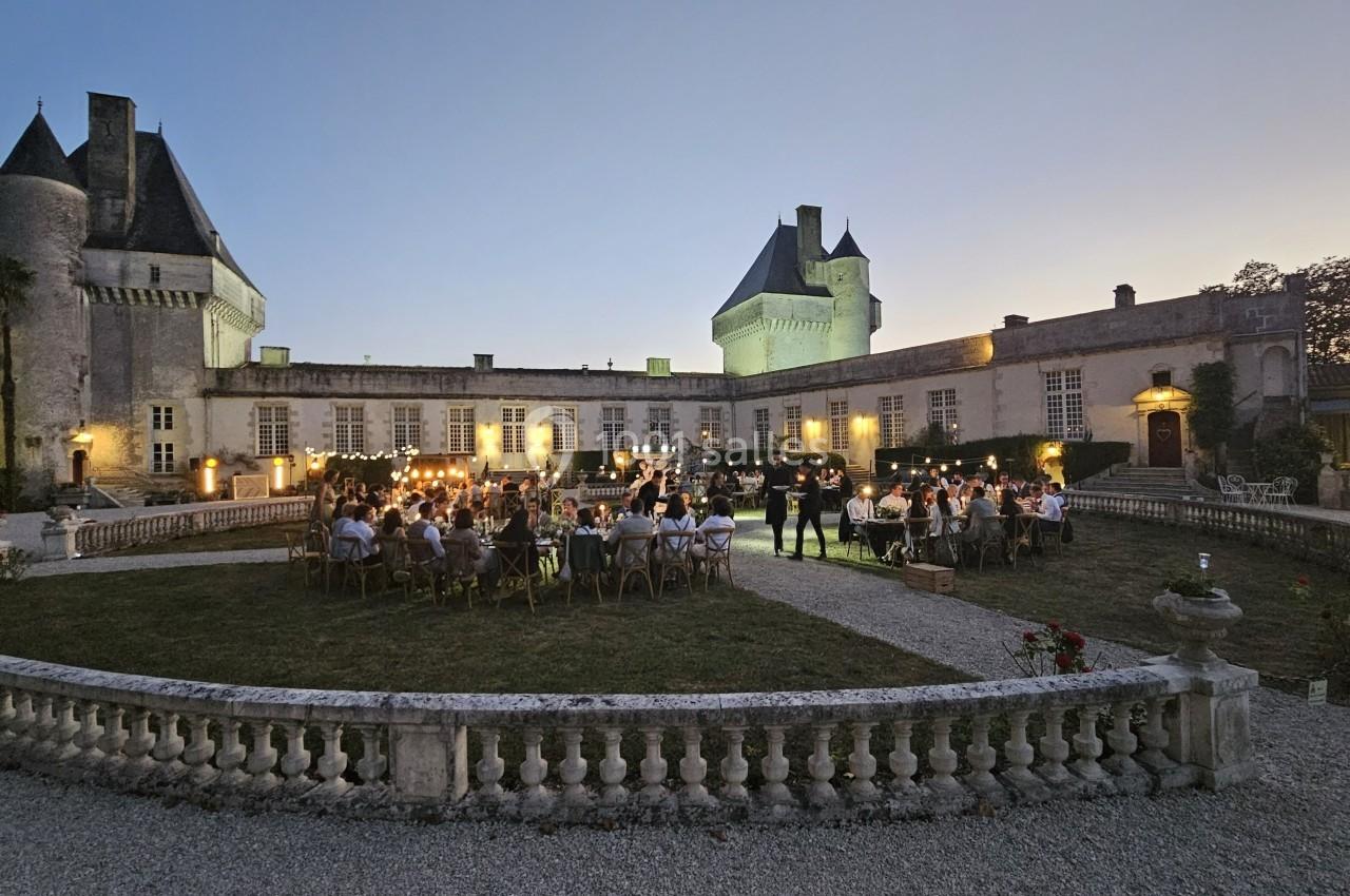 Dîner en plein air au crépuscule dans la cour d'un château illuminé, entouré de jardins et d'invités.