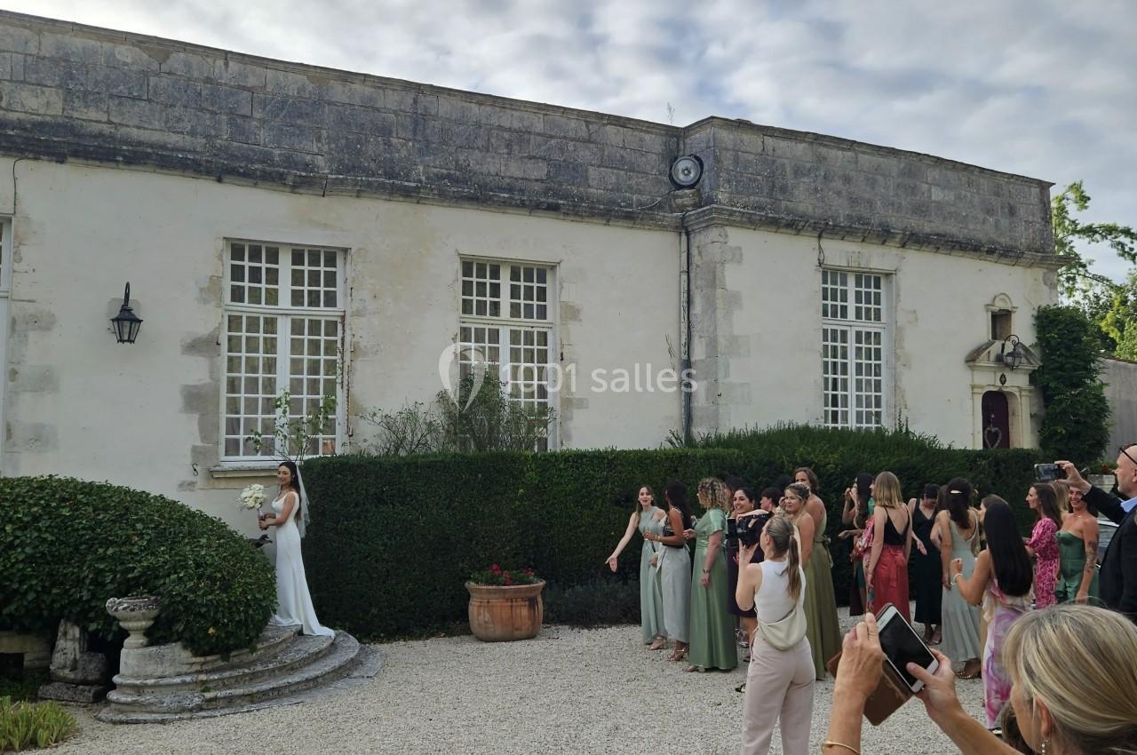 Un groupe de personnes observe une mariée tenant un bouquet devant un bâtiment ancien avec des fenêtres à carreaux.