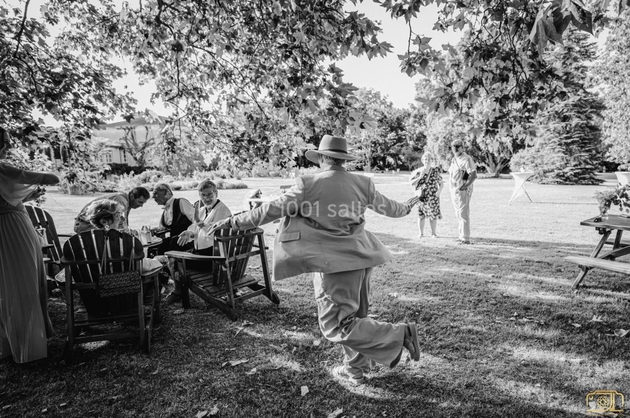 Un homme en costume clair danse joyeusement sur une pelouse, entouré de personnes assises sous des arbres.