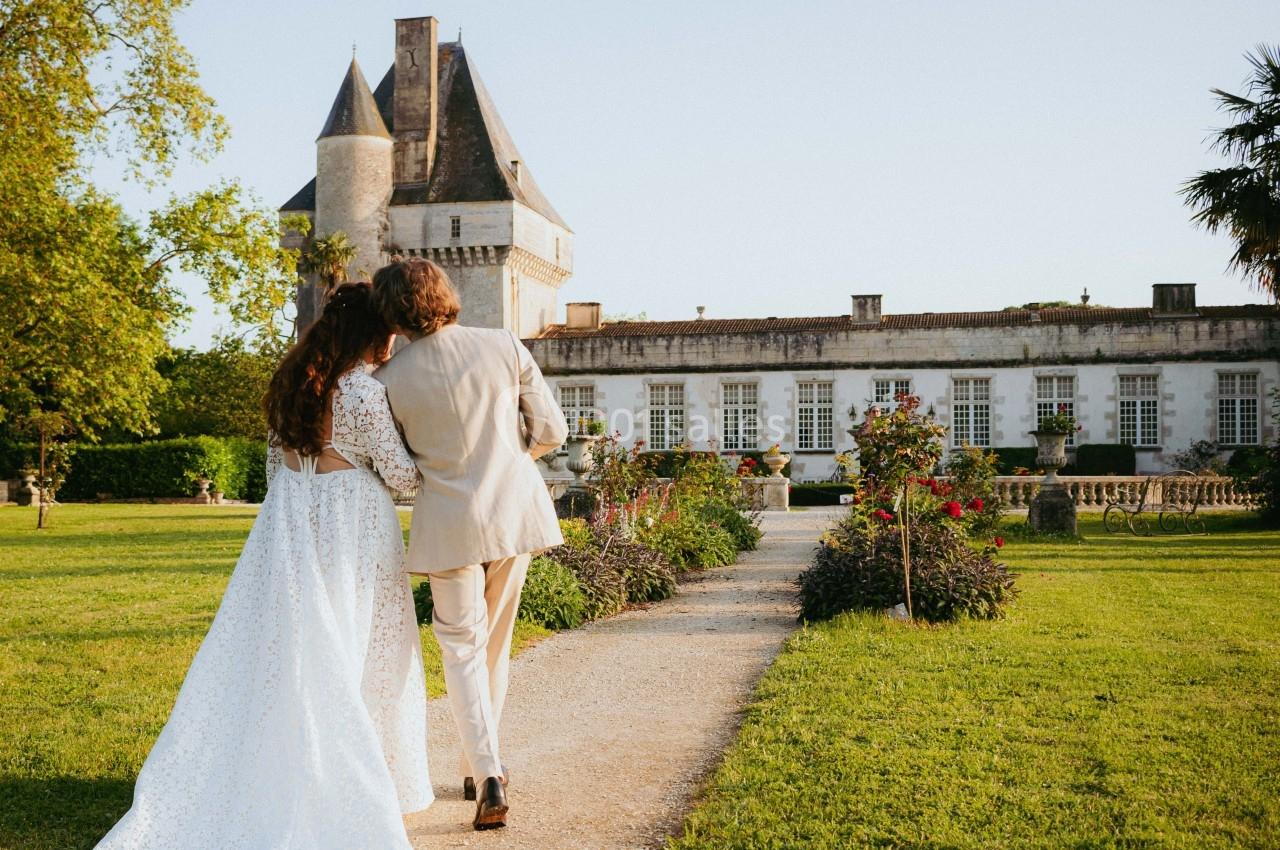 Un couple de dos marche sur un chemin bordé de verdure, menant à un château sous un ciel dégagé.