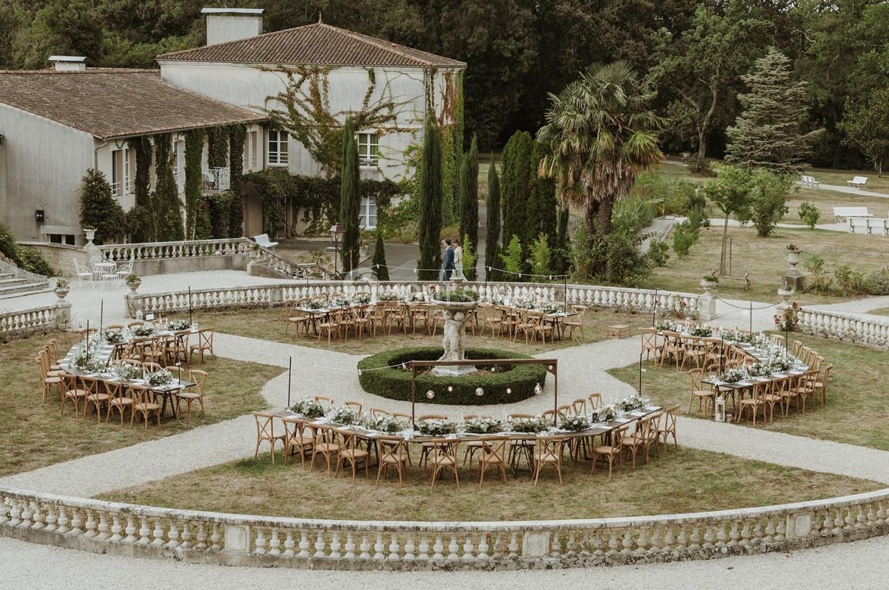 Tables dressées en cercle autour d'une fontaine dans un jardin, devant une maison de style traditionnel.
