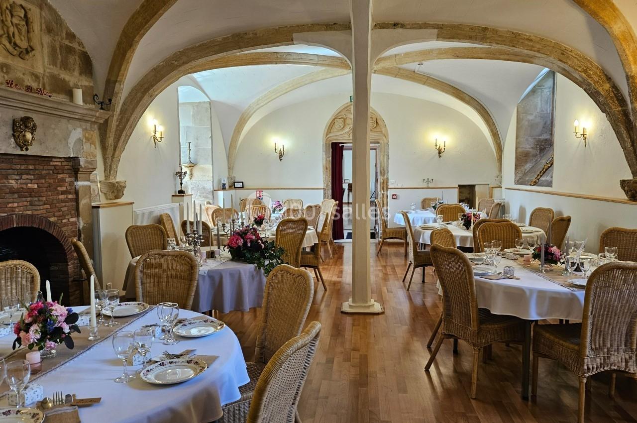 Salle de réception élégante avec tables dressées, chaises en osier, poutres apparentes et cheminée en pierre.