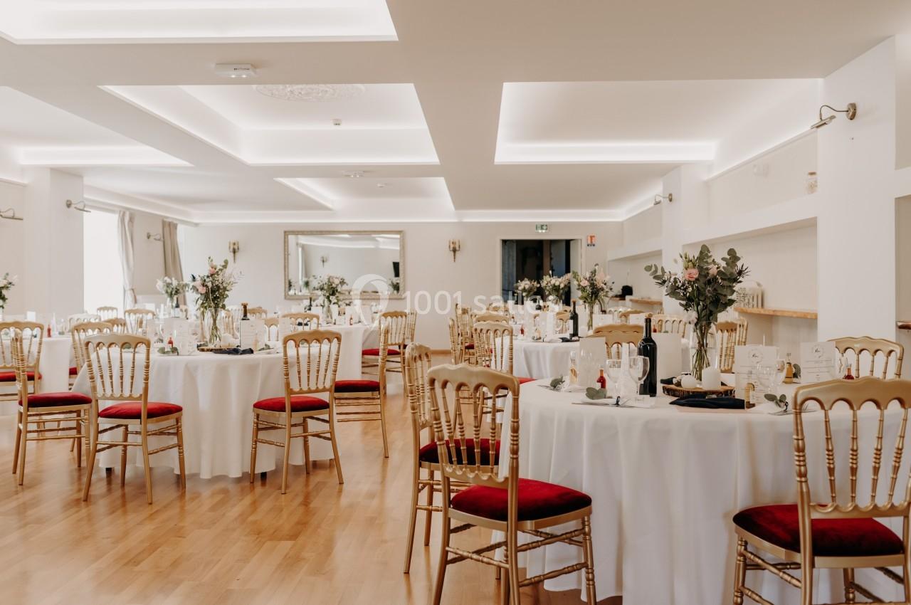 Salle de réception lumineuse avec tables rondes dressées, chaises dorées à assise rouge et décorations florales.