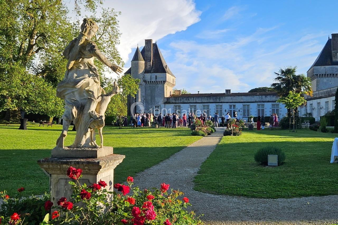 Statue en pierre dans un jardin fleuri, avec un château en arrière-plan et des personnes rassemblées au loin.