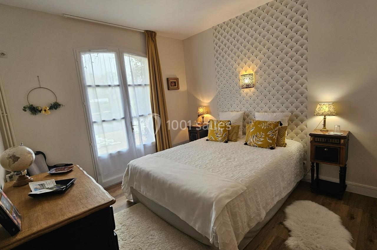 Chambre lumineuse avec lit double, coussins jaunes, mur capitonné, meubles en bois et fenêtre avec rideaux.