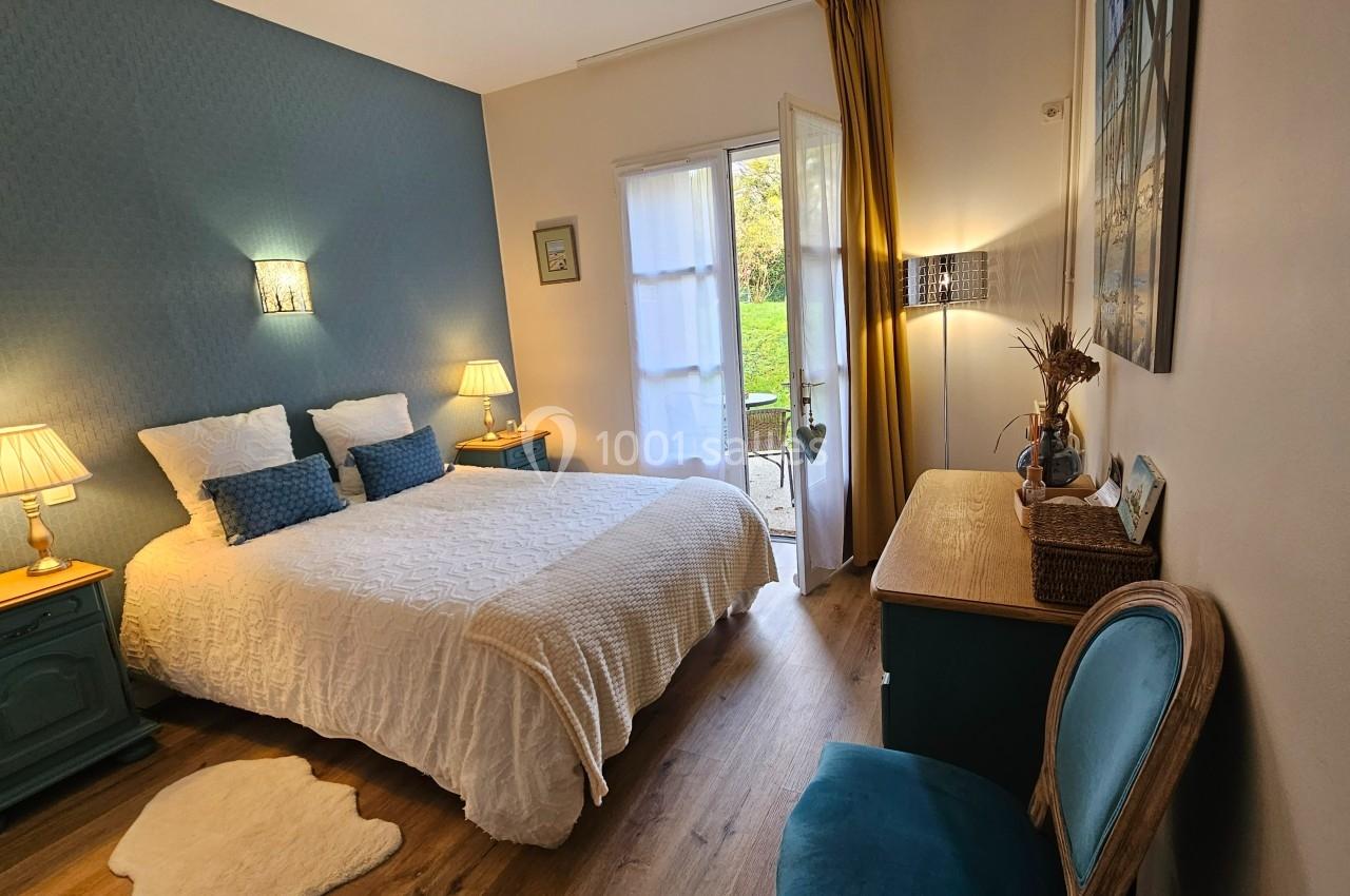 Chambre lumineuse avec lit double, murs bleus, mobilier en bois, et accès à une terrasse avec vue sur un jardin.