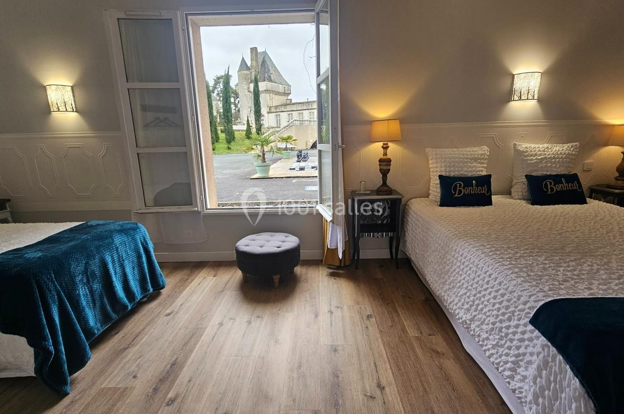 Chambre lumineuse avec deux lits, parquet, décoration élégante et vue sur un bâtiment historique par une fenêtre ouverte.