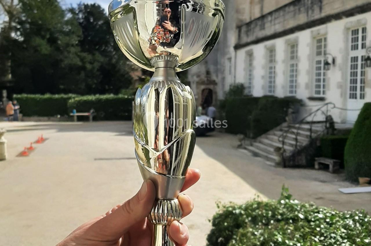 Une main tient un trophée devant un bâtiment historique entouré de verdure.