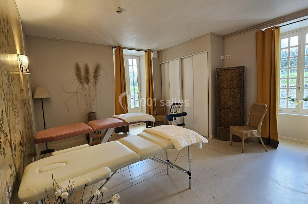 Salle lumineuse avec deux tables de massage, chaises en osier, décor floral et grandes fenêtres avec rideaux jaunes.