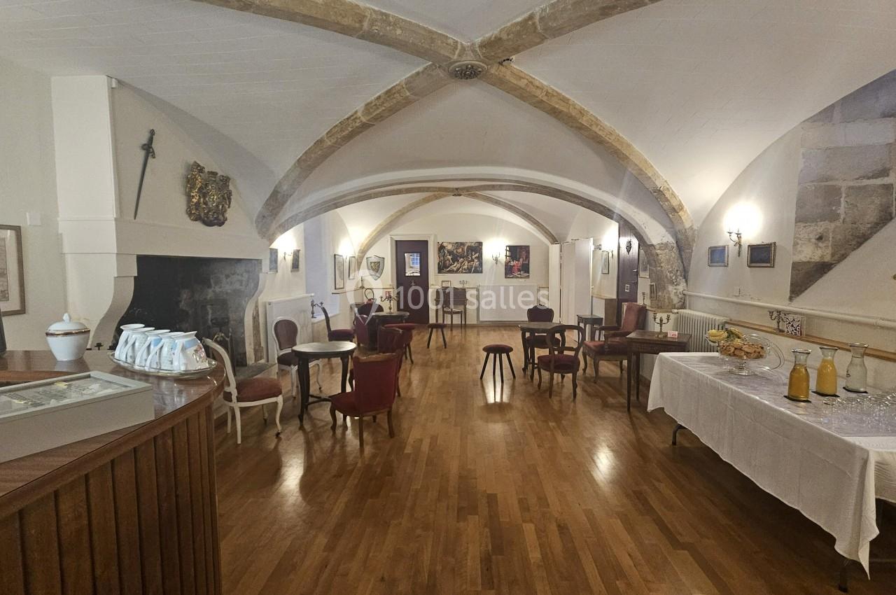 Salle voûtée avec parquet, tables et chaises, buffet dressé à droite et cheminée décorative à gauche.