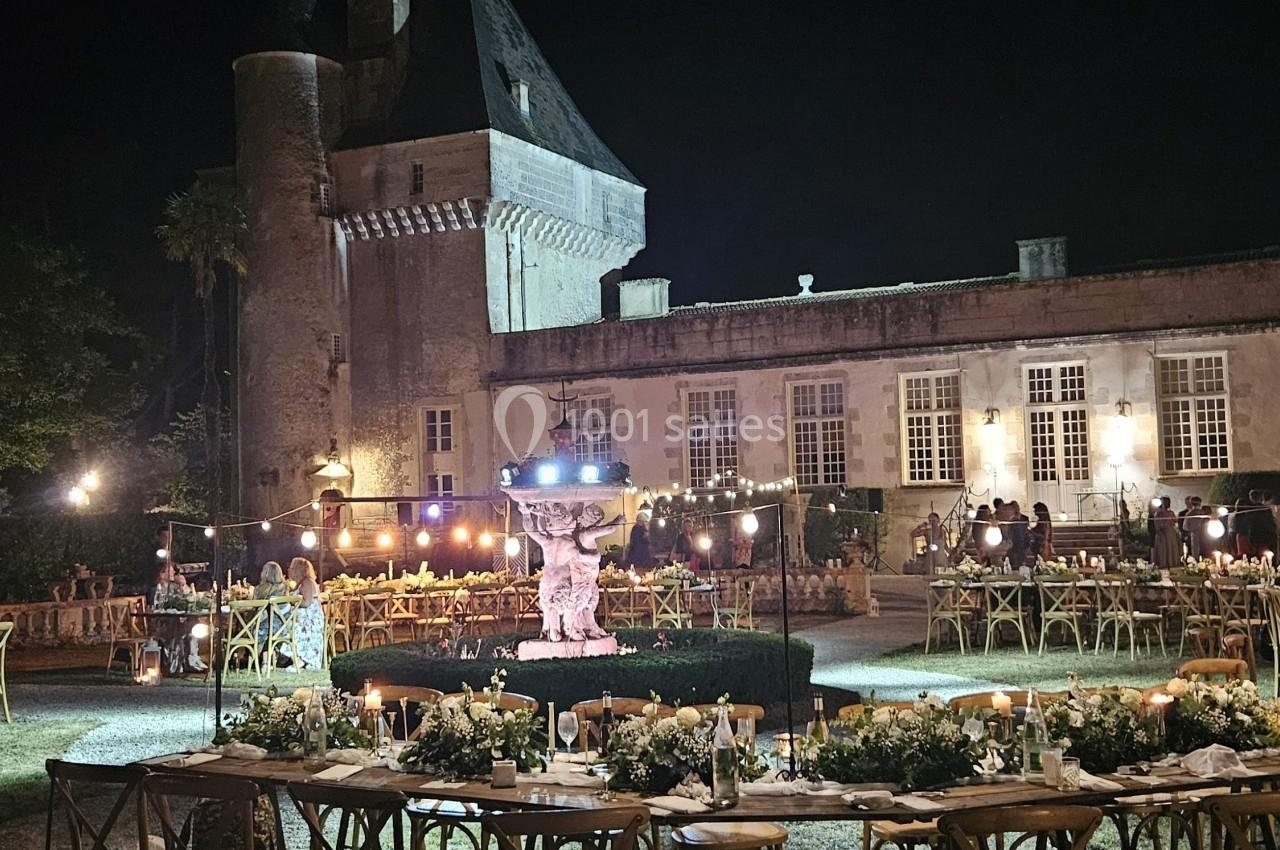 Vue nocturne d'un château éclairé, avec des tables décorées et des chaises disposées pour un événement en plein air.