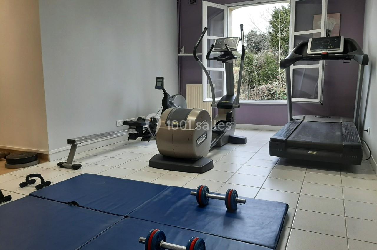 Salle de sport avec tapis de course, vélo elliptique, rameur, haltères et tapis au sol, près d'une fenêtre ouverte.