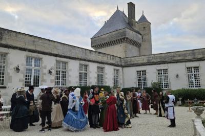 Des personnes en costumes d'époque discutent dans la cour d'un château historique sous un ciel partiellement nuageux.