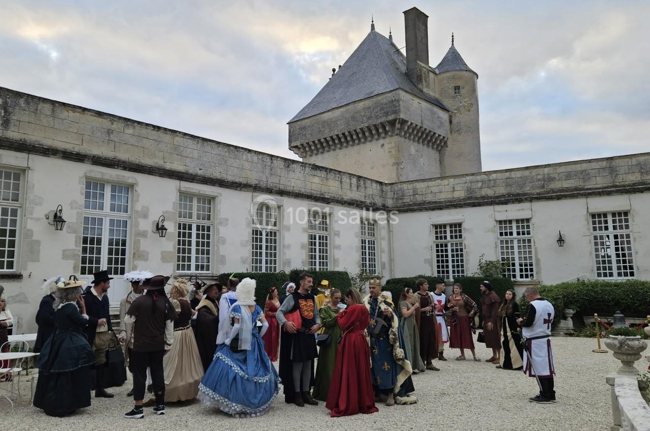 Des personnes en costumes d'époque discutent dans la cour d'un château historique sous un ciel partiellement nuageux.