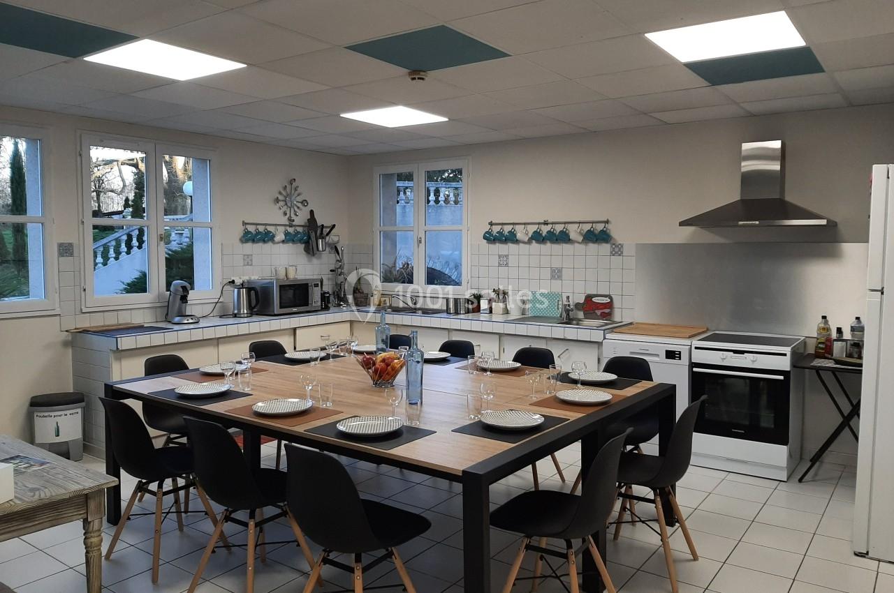 Cuisine spacieuse avec grande table dressée pour huit personnes, équipée d'appareils modernes et éclairée par des fenêtres.