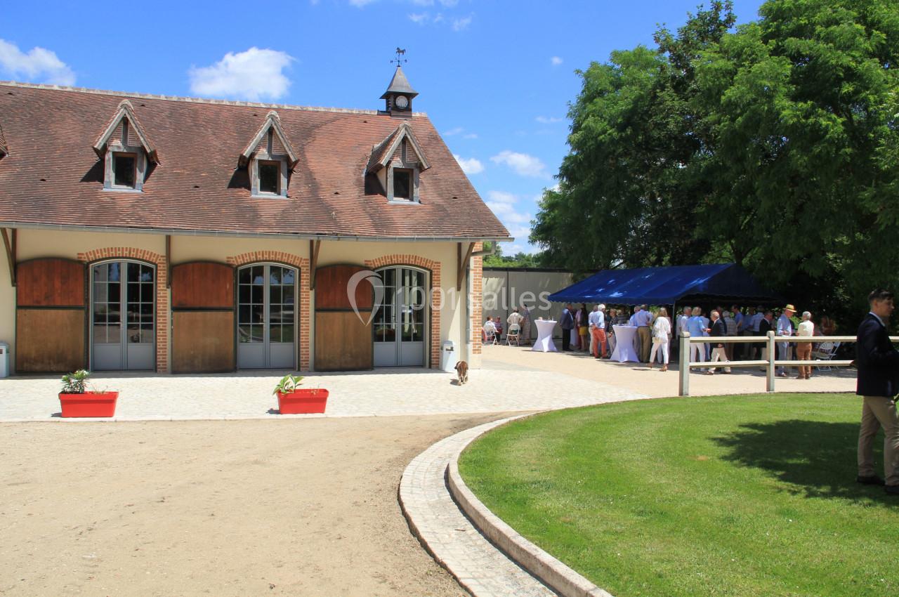 Bâtiment en briques avec toit en ardoise, terrasse pavée et groupe de personnes rassemblées sous un auvent bleu.
