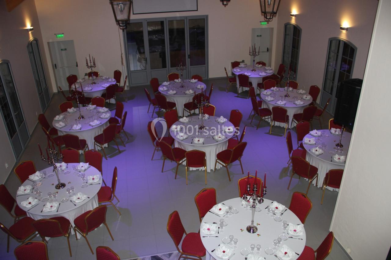 Salle de réception avec tables rondes dressées, nappes blanches, chaises rouges et chandeliers centraux.