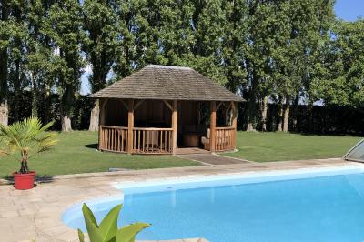 Piscine extérieure avec terrasse en pierre, cabane en bois et arbres en arrière-plan par une journée ensoleillée.