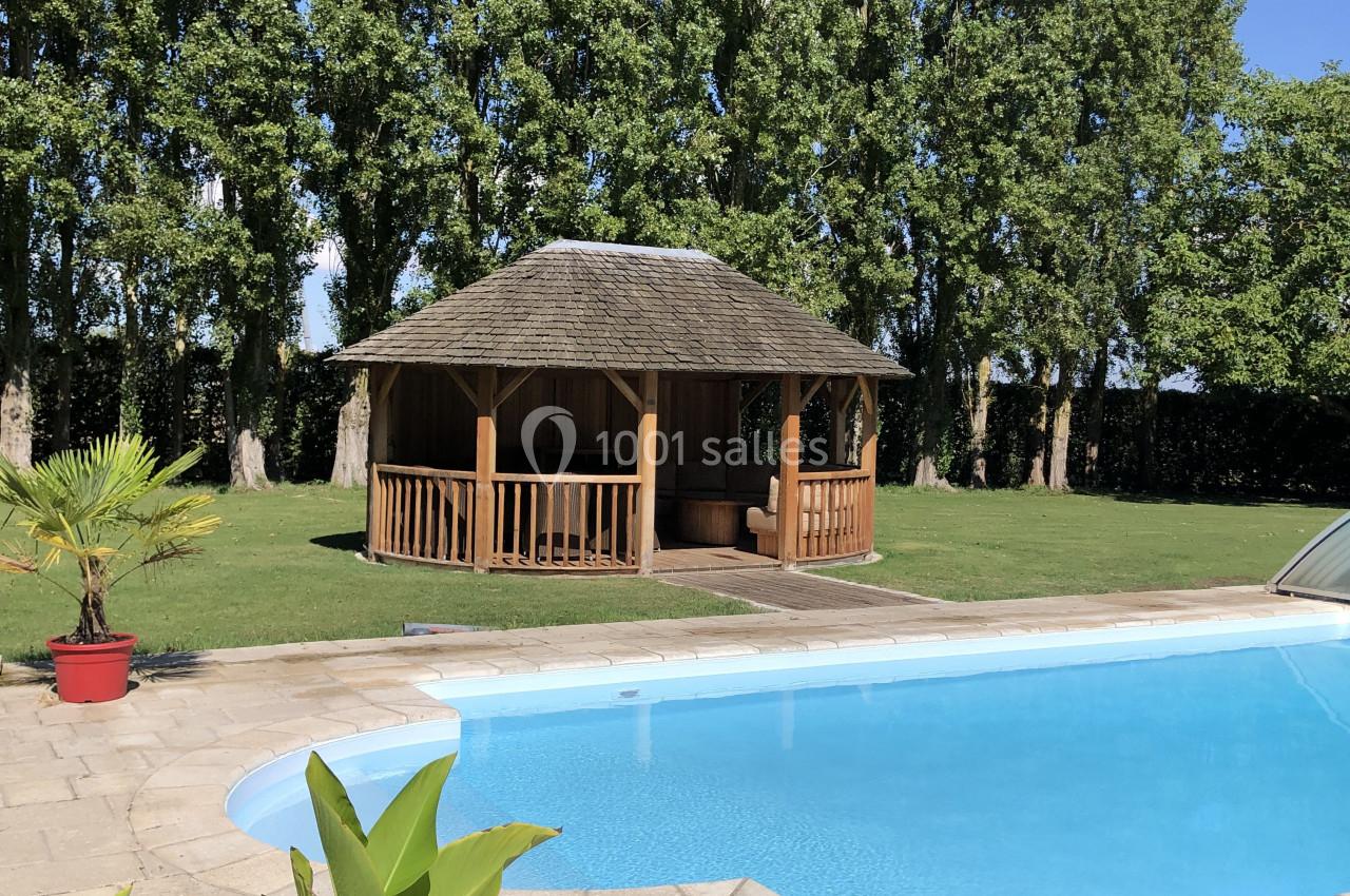 Piscine extérieure avec terrasse en pierre, cabane en bois et arbres en arrière-plan par une journée ensoleillée.