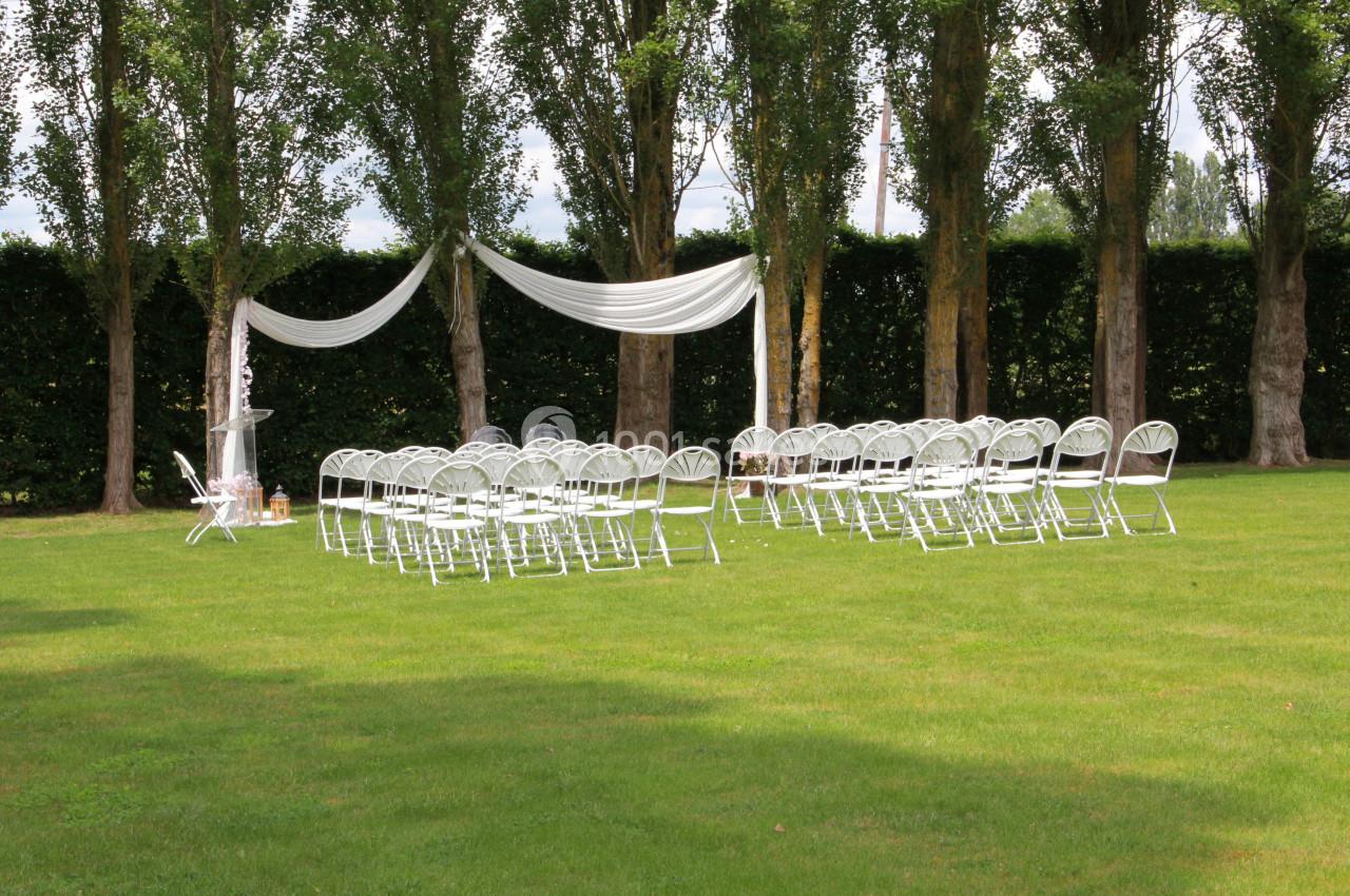 Chaises blanches disposées en rangées sur une pelouse, avec une arche décorée de tissu blanc devant des arbres.