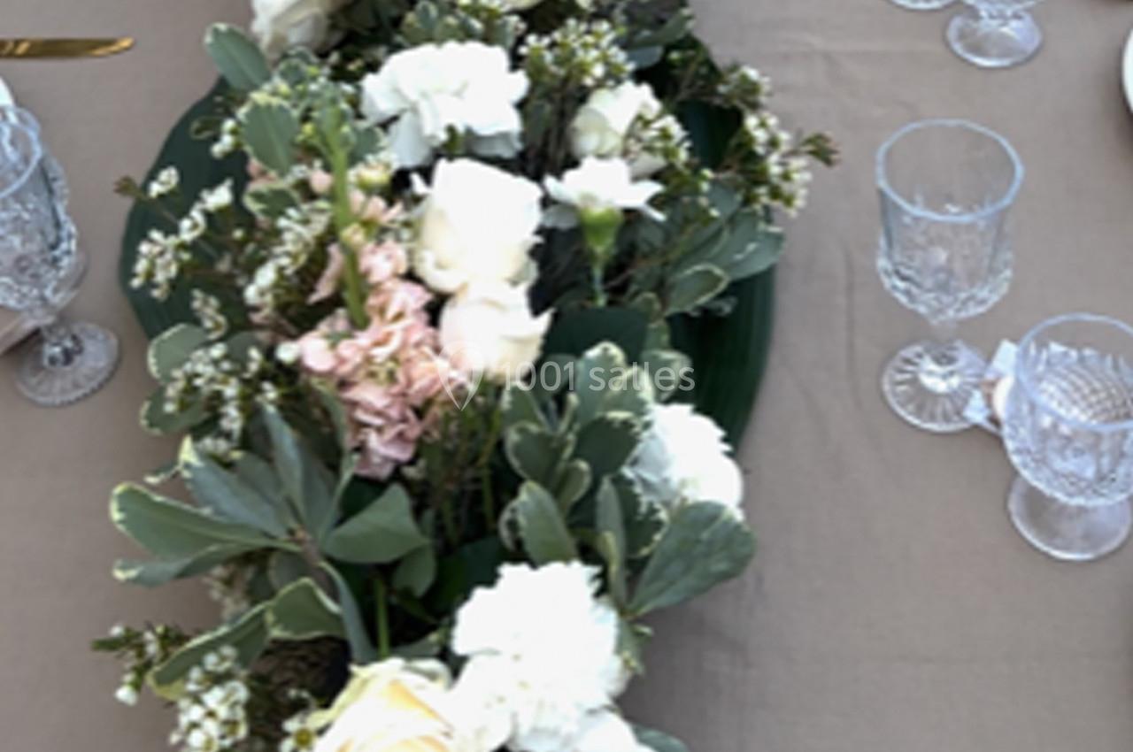 Centre de table floral avec des roses blanches, des fleurs roses et des feuillages verts, entouré de verres en cristal.