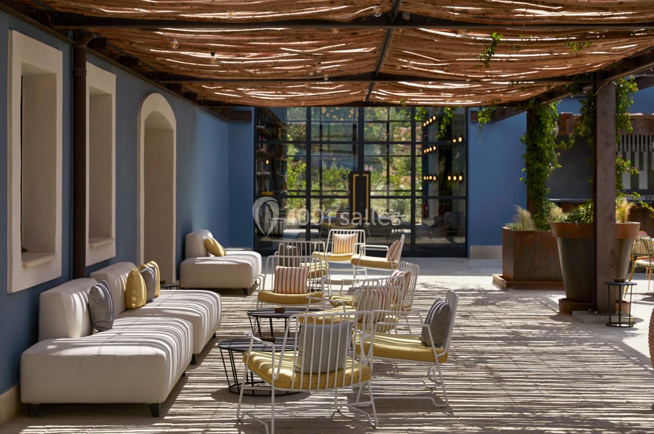 Terrasse ombragée avec mobilier moderne, canapés et chaises, entourée de plantes et d'une façade vitrée.