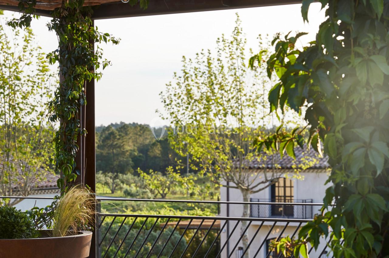 Table en bois dressée avec des verres et des lanternes sous une pergola en bois entourée de verdure, vue sur un paysage…