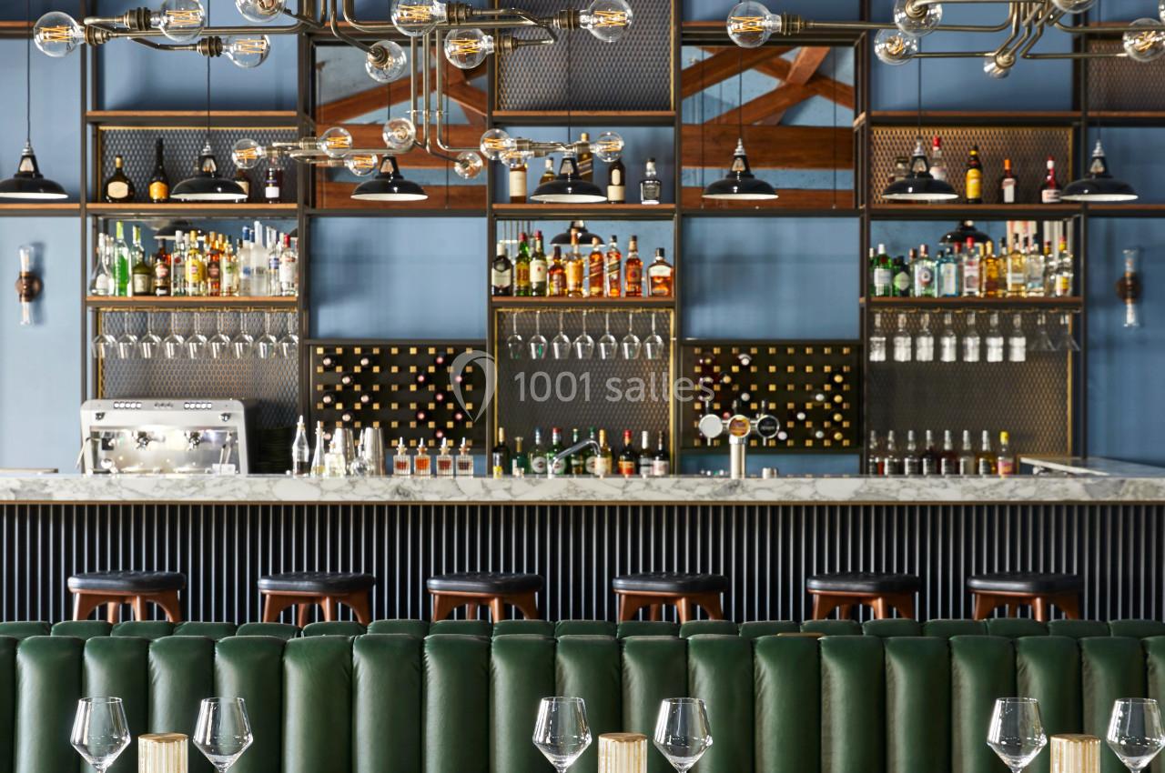 Bar élégant avec étagères remplies de bouteilles, banquettes vertes, tables dressées et luminaires suspendus modernes.