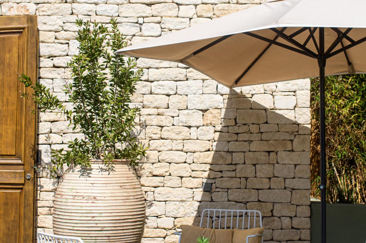 Mobilier de jardin avec fauteuils, table basse et parasol beige, disposés sur une terrasse en pierre devant un mur en pierre…