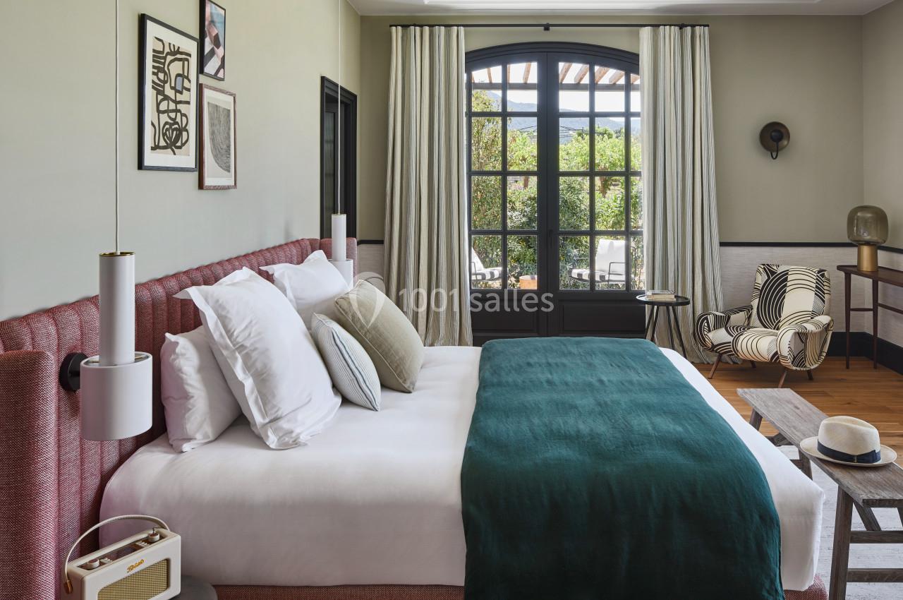 Chambre lumineuse avec lit double, coussins colorés, fauteuil rayé, grande fenêtre et vue sur un jardin.