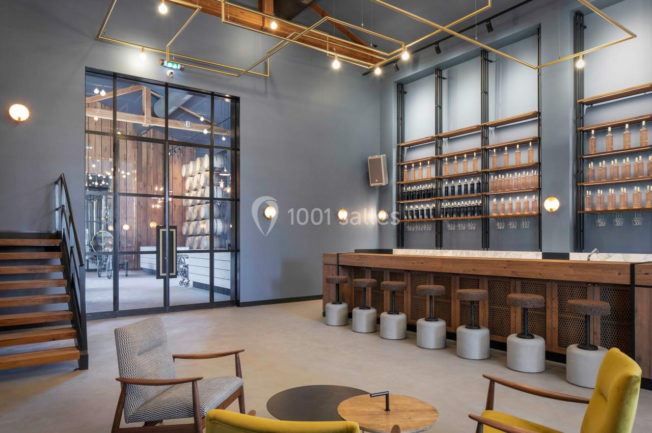 Salle moderne avec un bar en bois, tabourets hauts, fauteuils design et vue sur une pièce avec des fûts à travers une baie…