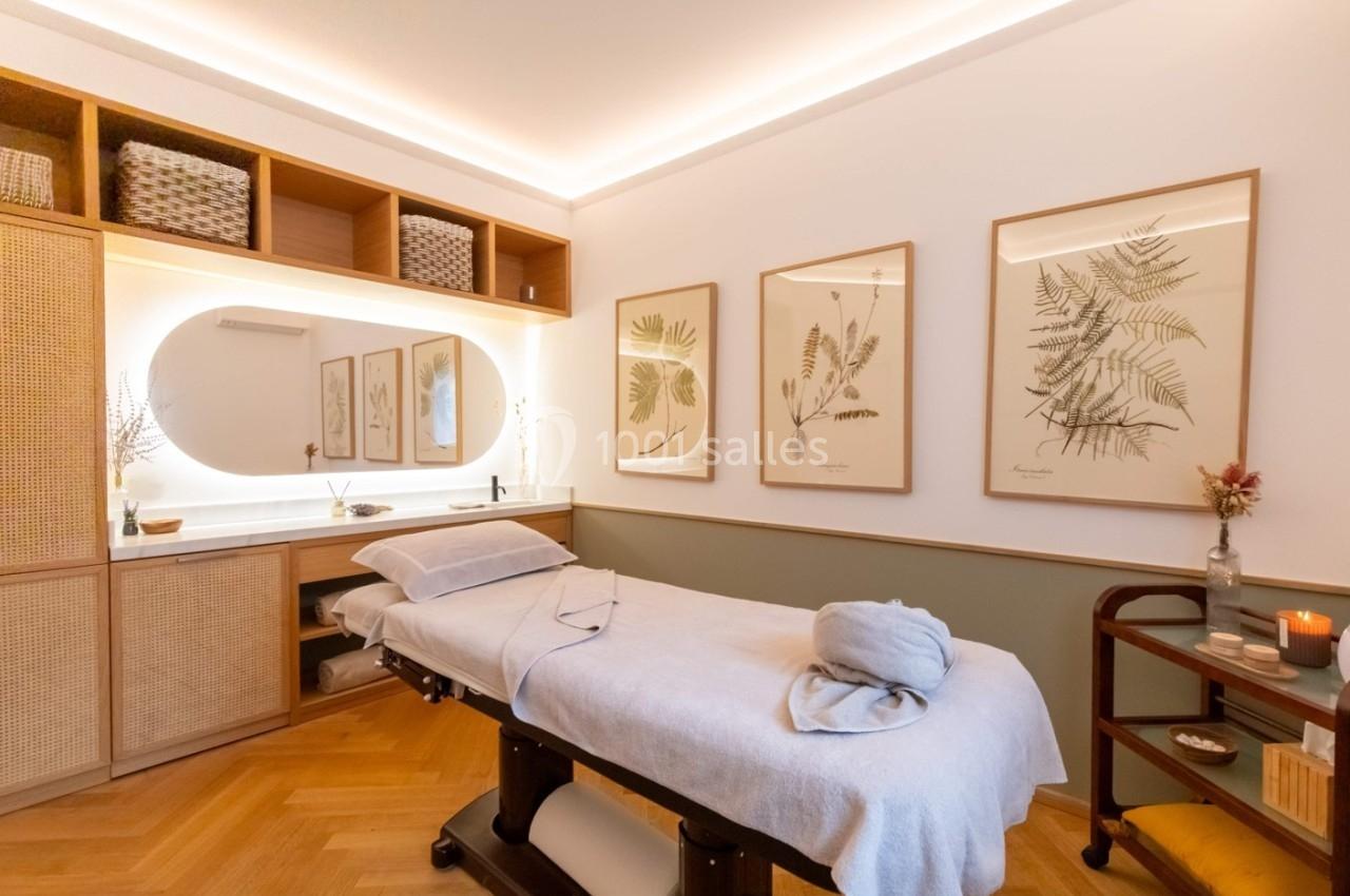 Salle de massage lumineuse avec table de soin, décorations murales végétales et éclairage chaleureux.