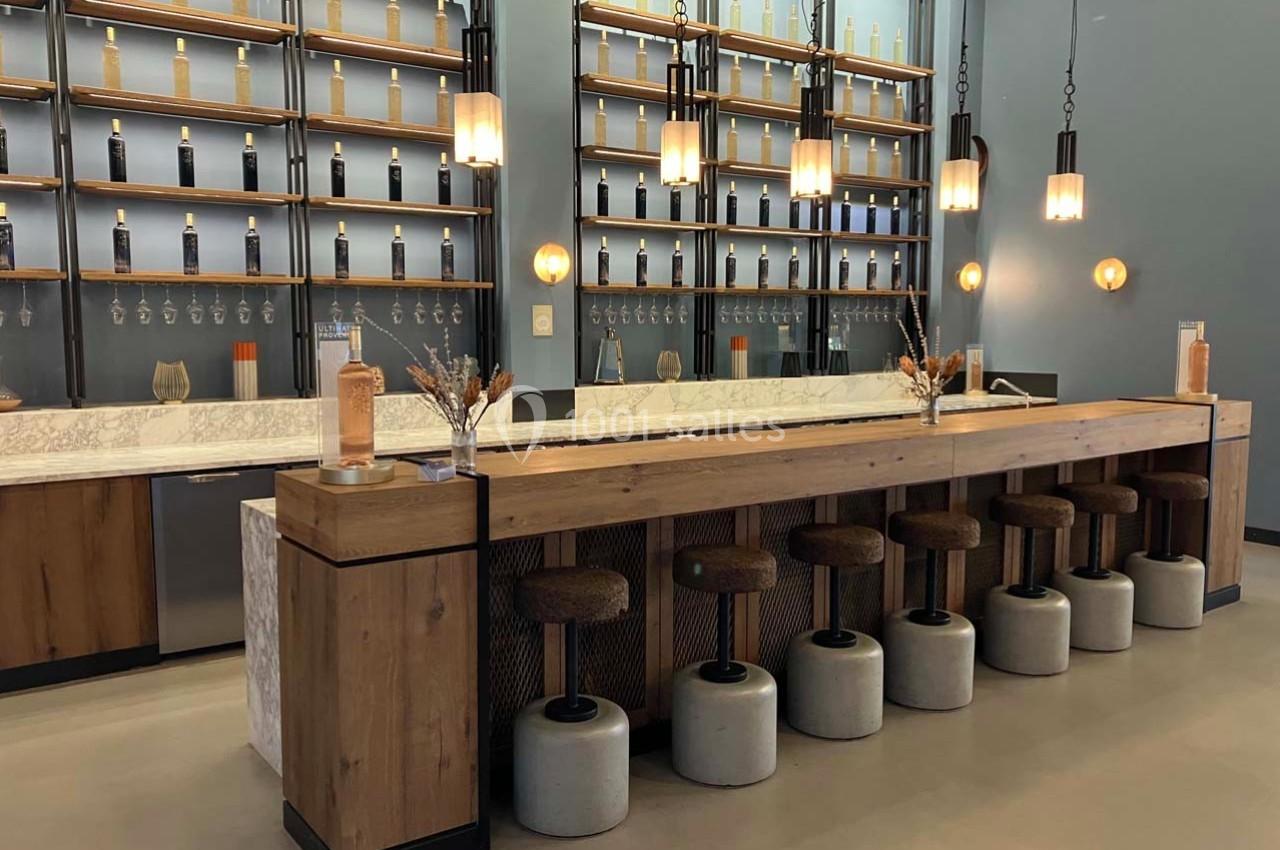 Bar moderne avec comptoir en bois, tabourets rembourrés, étagères murales garnies de bouteilles et éclairage suspendu.