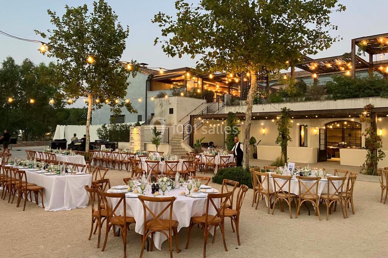 Tables rondes dressées avec nappes blanches dans un espace extérieur éclairé par des guirlandes lumineuses au crépuscule.