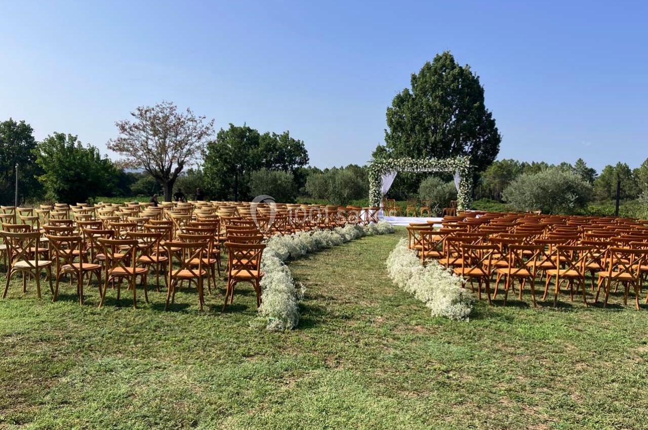 Chaises en bois disposées en rangées face à une arche fleurie dans un cadre extérieur verdoyant sous un ciel dégagé.