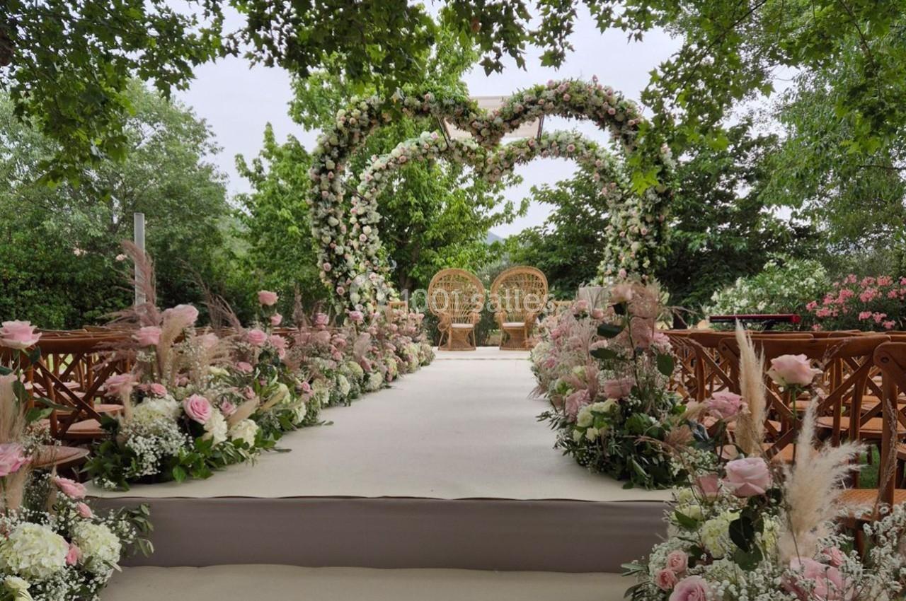 Allée décorée de fleurs menant à une arche florale en forme de cœur avec deux fauteuils en osier, dans un jardin verdoyant.
